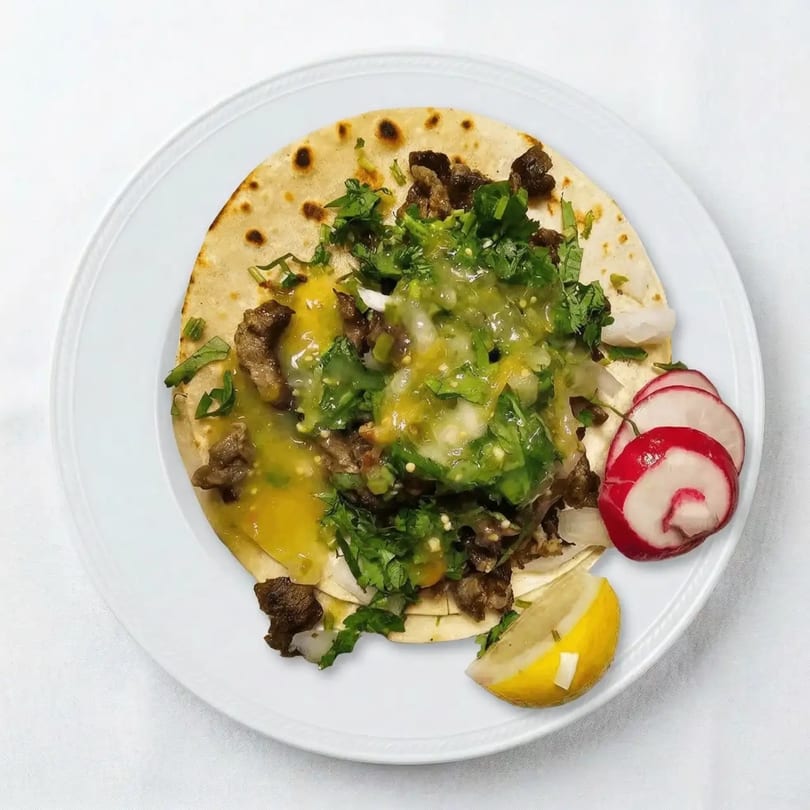 A single taco with meat, green and yellow salsa, cilantro, radishes, and a lemon wedge on a white plate.