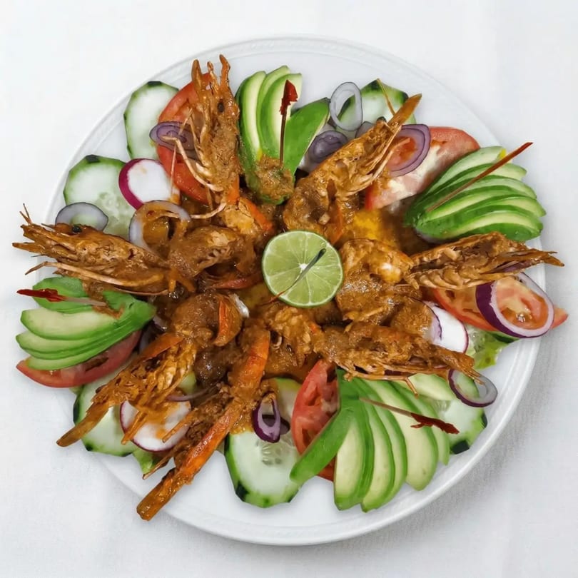 A plate of cooked shrimp with heads and tails, garnished with avocado, cucumber, tomato, and red onion.