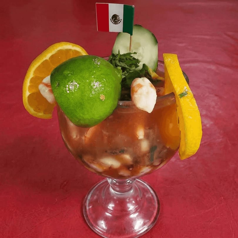 Mexican shrimp cocktail with lime, orange, cucumber, and a Mexican flag on a red background.